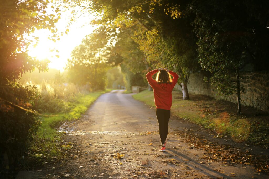 Person walking alone on a quiet tree-lined path at sunset, wearing a red sweater and black leggings, with arms resting on their head
