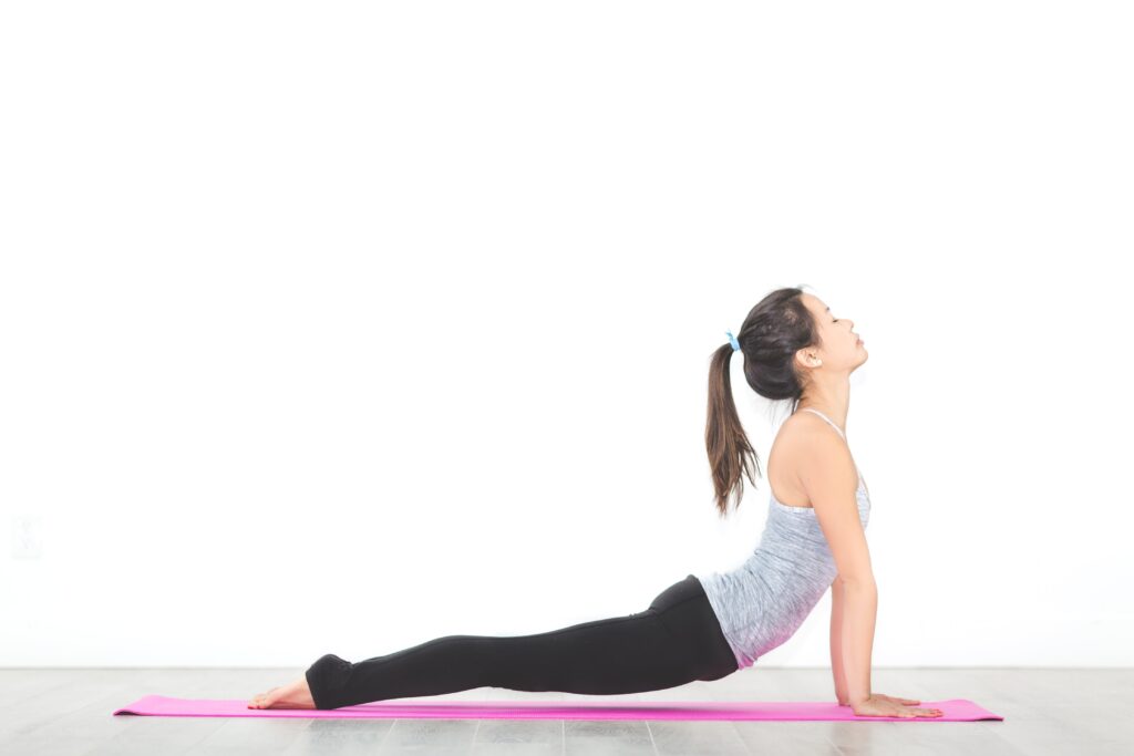 Woman practicing yoga indoors, performing an upward-facing stretch on a pink mat, wearing a gray tank top and black leggings