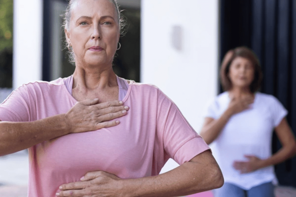 Two senior women practicing mindful breathing outdoors; the woman in the foreground wears a pink top with one hand on her chest and one on her abdomen, while another woman is softly blurred in the background doing the same pose.