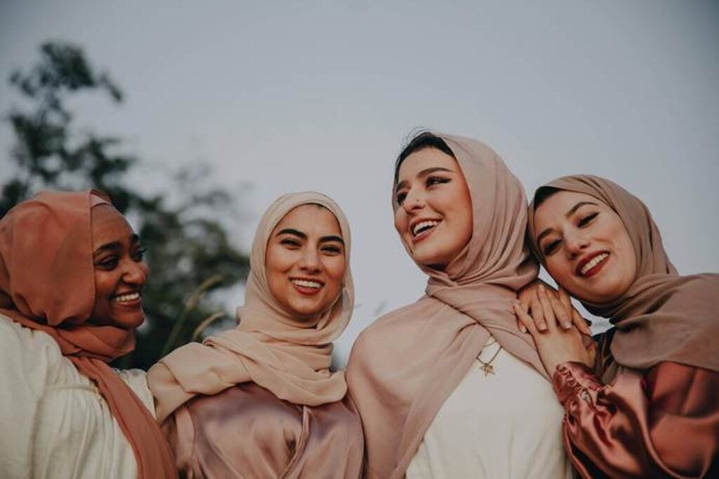 Four women wearing neutral-toned hijabs smiling and standing close together outdoors at golden hour, conveying friendship and support