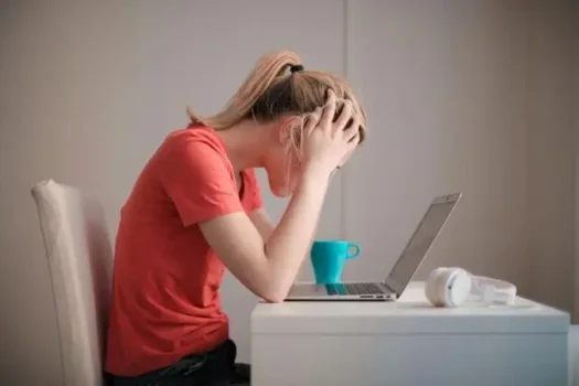 Woman in a red t-shirt sitting at a desk with her head in her hands, looking stressed in front of a laptop and coffee mug—representing the emotional and physical challenges of premenstrual syndrome (PMS).