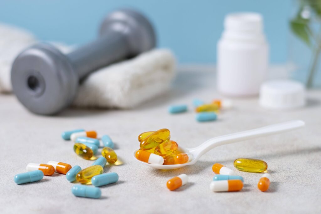 Close-up of various supplement capsules and softgels, including omega-3 and multivitamins, arranged on a spoon with a dumbbell and towel in the background—illustrating evidence-based supplements for PMS treatment.
