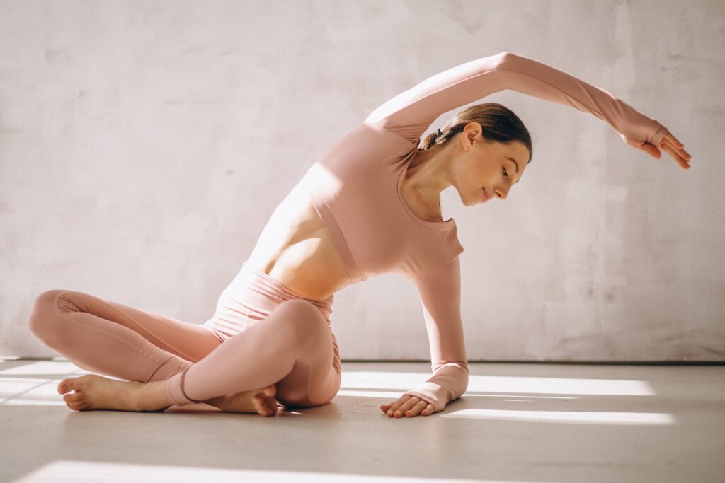 Young woman in pink activewear performing a seated side stretch on the floor in natural light—showcasing gentle movement and self-care as part of a PMS relief routine.