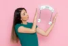 Woman in a teal t-shirt holding a transparent bathroom scale and looking at it against a pink background